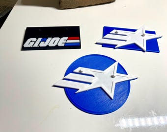Peut inclure: Trois objets imprimés en 3D : une plaque noire avec le texte « G.I. JOE » en lettres blanches avec des accents rouges et bleus, et deux plaques bleues avec une étoile blanche et trois bandes blanches.