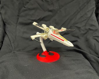 Puede incluir: Un modelo de caza estelar X-Wing blanco y rojo sobre una base roja con el símbolo de la Alianza Rebelde. El modelo tiene una cabina y alas detalladas.