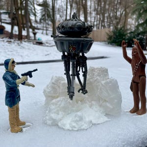 以下が含まれることがあります： 黒と灰色のスターウォーズのアクションフィギュア、歩いているAT-STと、その前に立っている茶色と白のチューバッカのアクションフィギュア。両方のフィギュアは白い雪の表面にあります。