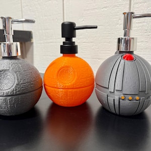 Puede incluir: Tres dispensadores de jabón esféricos con mecanismos de bomba. El de la izquierda es gris oscuro con superficie texturizada, el central es naranja brillante con bomba negra y el de la derecha es gris claro con un detalle rojo y tres puntos amarillos.