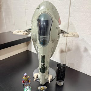 May include: A gray toy spaceship with a central cockpit and wing-like extensions. A small Boba Fett action figure, a small gray and orange object, and a black rectangular object are in front of the spaceship.