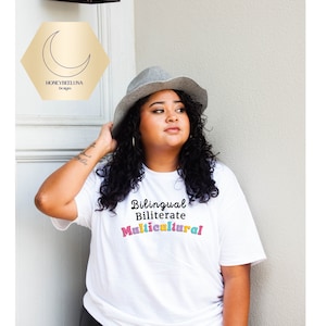 May include: A white short-sleeved t-shirt featuring the black text Bilingual, Biliterate and the word Multicultural in colorful, bubble-style letters. The shirt is paired with a grey fedora hat, and a logo with a crescent moon and the text HONEYBEELUNA Designs.