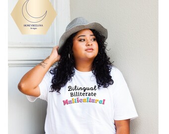 May include: A white short-sleeved t-shirt featuring the black text Bilingual, Biliterate and the word Multicultural in colourful, bubble-style letters. The shirt is paired with a grey fedora hat, and a logo with a crescent moon and the text HONEYBEELUNA Designs.