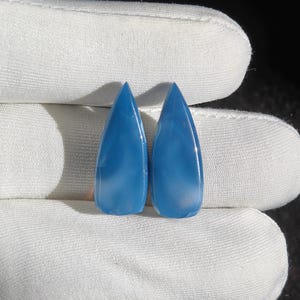 May include: Two teardrop-shaped, polished blue gemstones. The stones are a vibrant, solid blue color with a smooth, glossy surface. The gemstones are displayed against a white background.
