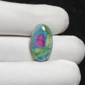May include: An oval gemstone with a striking colour palette. The centre displays a rich magenta, encircled by shades of teal, green, and yellow. The polished surface reflects light, highlighting the stone's natural patterns and textures.