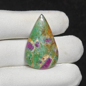 Puede incluir: Una piedra preciosa en forma de lágrima con una superficie pulida. La piedra presenta una mezcla de colores, incluyendo verde, rosa y naranja. El verde es el color dominante, con parches rosas y naranjas. La piedra es probablemente un colgante o cabujón.