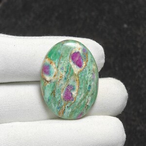May include: Oval-shaped gemstone with a green base color, featuring vibrant pink and purple inclusions. The stone has a polished surface, and the inclusions are irregularly shaped. The gemstone is held in a white fabric.