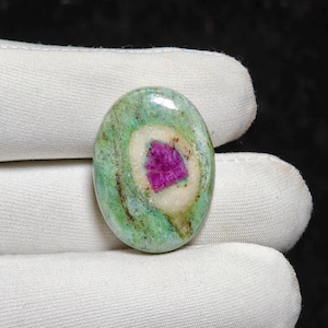 Könnte beinhalten: Ovalförmiger Edelstein mit lebendiger Farbpalette. Der Stein hat einen zentralen, tiefmagentafarbenen Kern, der von einem weißen Band und einer gesprenkelten grünen Außenschicht umgeben ist. Die polierte Oberfläche reflektiert das Licht.