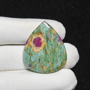 May include: A teardrop-shaped gemstone with a polished surface. The stone features a vibrant mix of colors, including green, red, and yellow. The red areas are concentrated in the center and edges, while the green and yellow hues create a marbled effect.