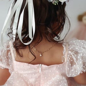 May include: A young person wearing a light pink dress with white polka dots and a large bow in the back. The dress has sheer, puffy sleeves. A gold necklace with a small pearl pendant is visible. The person is wearing a floral crown and white ribbons in their hair.