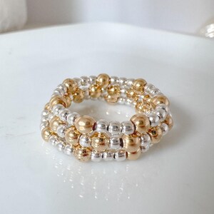 May include: A gold and silver beaded ring. The ring features three rows of small silver beads and larger gold beads. The ring is on a white surface.