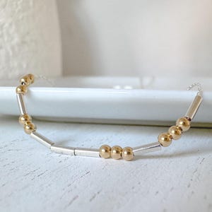 Könnte beinhalten: Ein zartes Armband mit einem abwechselnden Muster aus kleinen, runden goldfarbenen Perlen und schlanken, zylindrischen silberfarbenen Metallröhrchen, die auf einer dünnen, flexiblen Kette aufgereiht sind und eine raffinierte Optik bieten.