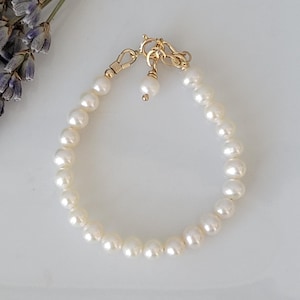 Könnte beinhalten: Ein zartes Perlenarmband mit einem goldenen Verschluss und Akzenten. Das Armband besteht aus runden, cremeweißen Perlen. Ein Zweig getrockneten Lavendels befindet sich in der oberen linken Ecke auf weißem Hintergrund.