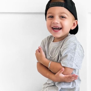 May include: A young child wearing a gray t-shirt, black baseball cap, and black pants. The child is also wearing a silver chain necklace and bracelet. The t-shirt has the initials "LG" on the side. The child is wearing white and black sneakers.