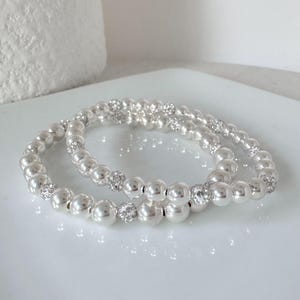 Puede incluir: Dos pulseras de cuentas plateadas. Cada pulsera presenta cuentas plateadas grandes, redondas y brillantes, intercaladas con cuentas más pequeñas y brillantes con incrustaciones de cristal. Las pulseras se exhiben sobre una superficie blanca y reflectante.