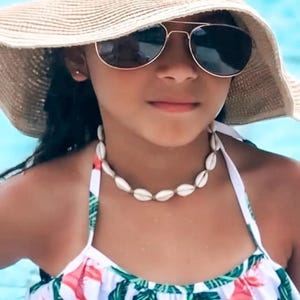 May include: A young person wearing a straw sun hat, aviator sunglasses, and a seashell necklace. The person is also wearing a white and floral print swimsuit. The background is a blurred blue.