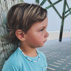 May include: A child wearing a necklace made of white and blue beads. The necklace is around the child's neck. The child is wearing a light blue t-shirt. The background shows a wooden post and a beach scene.