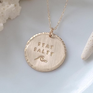 May include: Gold-colored pendant necklace with the words "STAY SALTY" and a wave design. The pendant is round with a textured surface and hangs from a delicate chain. The necklace is displayed on a light blue surface.