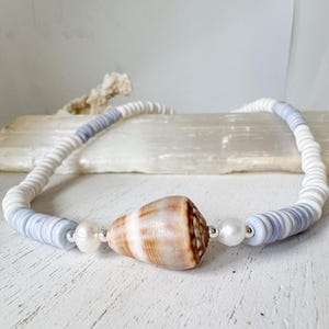 May include: A necklace with a large, brown and white seashell pendant. The necklace is made of white and light blue disc-shaped beads, with a pearl accent on each side of the shell. The necklace is on a white surface.