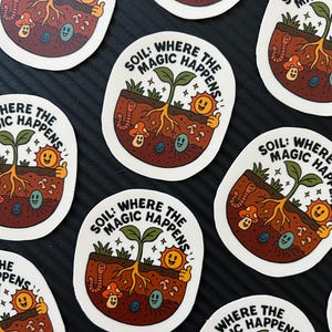 以下が含まれることがあります： 「SOIL: WHERE THE MAGIC HAPPENS」の文字が入った楕円形のステッカー。土、根、植物、太陽、ミミズ、キノコの漫画イラストが描かれています。ステッカーは白で、背景は黒です。