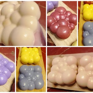 May include: A selection of handmade soaps in shades of white, pink, purple, yellow, and blue. Each soap is crafted to resemble a cluster of bubbles and is presented on a wooden board. The soaps have a shiny appearance.