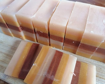 Puede incluir: Un juego de barras de jabón hechas a mano con capas de marrón, naranja y blanco. Las barras de jabón están dispuestas en una fila sobre una superficie de madera.