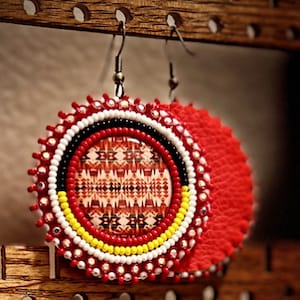 May include: A pair of red and white beaded earrings with a geometric pattern in the center. The earrings are hanging from a wooden display stand.