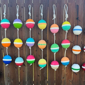 May include: A wooden fence decorated with a string of colorful, round, painted buoys. The buoys are hung with white rope and have various patterns and colors, including red, blue, green, yellow, and pink.