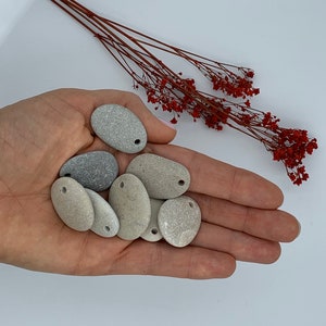 May include: A collection of smooth, gray and beige stones with holes drilled through them. The stones are arranged in a hand, with the holes facing up.