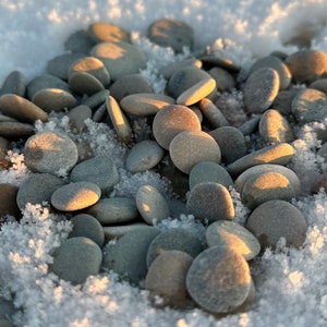 May include: A collection of smooth, gray stones arranged in a pile on a bed of white snow. The stones are various sizes and shapes, with some showing subtle variations in color.