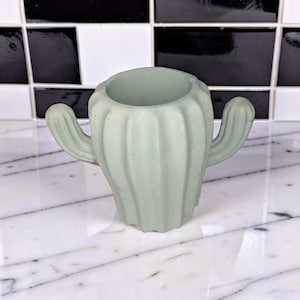 May include: A light sage green cactus-shaped container with a smooth, matte finish. The vessel features a rounded central body with vertical ribbed detailing and two small, curved arms extending from the sides, resembling a saguaro cactus.