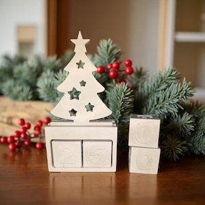 Puede incluir: Un calendario perpetuo blanco en forma de árbol de Navidad con recortes de estrellas, que muestra la fecha 25. Dos bloques adicionales muestran los números 0 y 0. El calendario está sobre ramas de hoja perenne y bayas rojas.