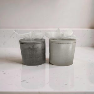 Puede incluir: Dos cajas de pañuelos de hormigón, una gris oscuro y otra gris claro, están una al lado de la otra. Cada una tiene una tapa plana y un paquete de pañuelos blancos que sobresale de la parte superior. Los soportes son cilíndricos y están sobre una superficie blanca reflectante.
