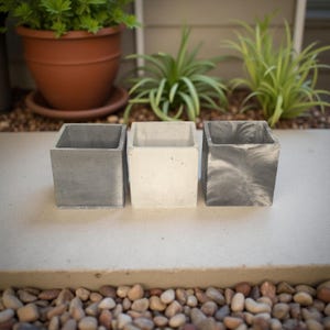 May include: Three square concrete planters in shades of gray and white are arranged in a row. The planters have a modern, minimalist design, perfect for succulents or small plants. The background includes a terracotta pot and greenery.