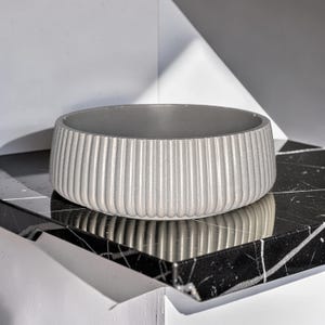 May include: A round, light gray bowl featuring a vertical ribbed texture along its exterior wall. The bowl has a smooth, flat rim and a consistent matte finish, presenting a simple, cylindrical shape with a hollow interior and a solid base.