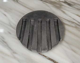 May include: A round, grey concrete coaster with five vertical grooves. The coaster is on a white marble surface.