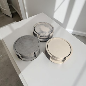 May include: Three sets of round concrete coasters. Each set has four coasters stacked on top of each other. The coasters are in three different colors: gray, white, and a marbled gray and white.