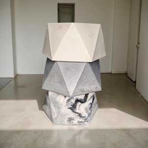 Könnte beinhalten: Drei geometrische Beton-Pflanzkübel, die übereinander gestapelt sind. Der obere Kübel ist weiß, der mittlere Kübel ist grau und der untere Kübel ist marmoriert in Grau und Schwarz.