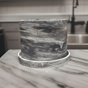 May include: A gray and white marbled ceramic planter with a matching saucer. The planter has a smooth, rounded shape and a subtle swirl pattern.