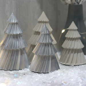 May include: A display of decorative Christmas trees in silver and white. The trees feature a layered, pleated design and are set on artificial snow. A string of fairy lights provides a warm glow to the festive scene, creating a wintery atmosphere.