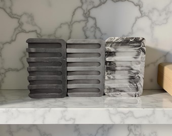May include: Three rectangular soap dishes made of gray and white speckled silicone. The dishes have raised ridges on the bottom to help drain water.