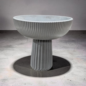 May include: A light gray, ribbed concrete pedestal bowl. The bowl has a wide, shallow design with vertical grooves. It sits atop a matching, fluted pedestal base. The bowl casts a dark shadow on the concrete floor.