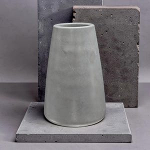 May include: A light gray, tapered cylindrical vase with a smooth, matte finish. The vase features a wide base that narrows toward the top opening, showcasing a solid, uniform color and a clean, simple structure without any additional patterns or designs.