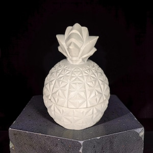 Peut inclure: Un récipient gris clair en forme d'ananas avec un motif géométrique en losanges texturé sur sa surface. L'objet présente un couvercle amovible surmonté d'une structure de feuilles pointues stylisées, fabriqué dans un matériau solide et mat.