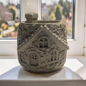 Puede incluir: Una maceta gris en forma de casa con chimenea, ventanas y una puerta. El diseño detallado incluye un techo y paredes texturizadas, creando una apariencia de cabaña en miniatura. La maceta está sobre un alféizar blanco.