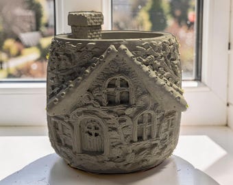 May include: A gray, house-shaped planter with a chimney, windows, and a door. The detailed design includes a textured roof and walls, creating a miniature cottage appearance. The planter sits on a white windowsill.