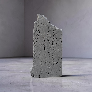 May include: A gray, rectangular concrete block with a rough, porous texture and a jagged edge. The block stands upright on a gray surface, showcasing its industrial aesthetic. The block's dimensions are approximately 12 inches tall and 6 inches wide.