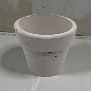 May include: A small, unglazed concrete flower pot with a slightly textured surface. The pot is light gray with a cylindrical shape and a wider rim. The interior is smooth and empty, ready for planting.