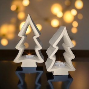 May include: Two off-white, tree-shaped candle holders. Each holder has a cutout design and a small space to hold a tea light candle. The holders are on a dark surface with a blurred background of warm, out-of-focus lights.