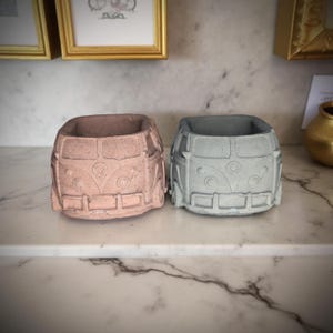 May include: Two small concrete planters shaped like vintage Volkswagen vans. One planter is painted a light pink color and the other is painted a light blue-gray color.